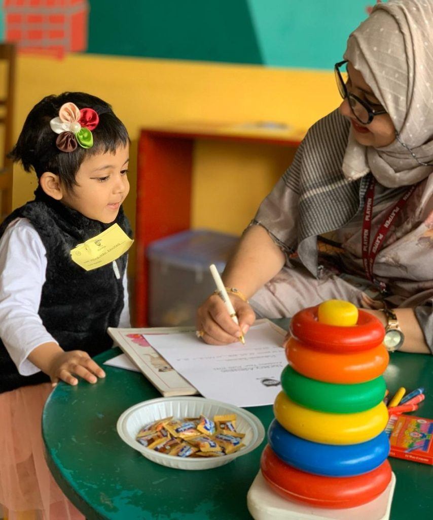 A young Playpen pupil writing at her desk with a teacher